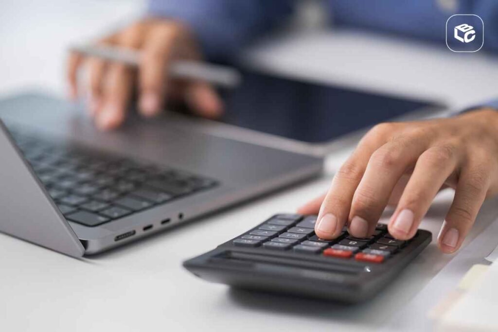 Close-up de uma pessoa utilizando uma calculadora sobre uma mesa, enquanto trabalha em um notebook ao fundo. Uma das mãos pressiona as teclas da calculadora e a outra segura uma caneta próxima ao computador. A imagem tem foco nos equipamentos de trabalho e remete a atividades de cálculo, finanças ou planejamento administrativo. Forneça seus comentários sobre o BizChat
